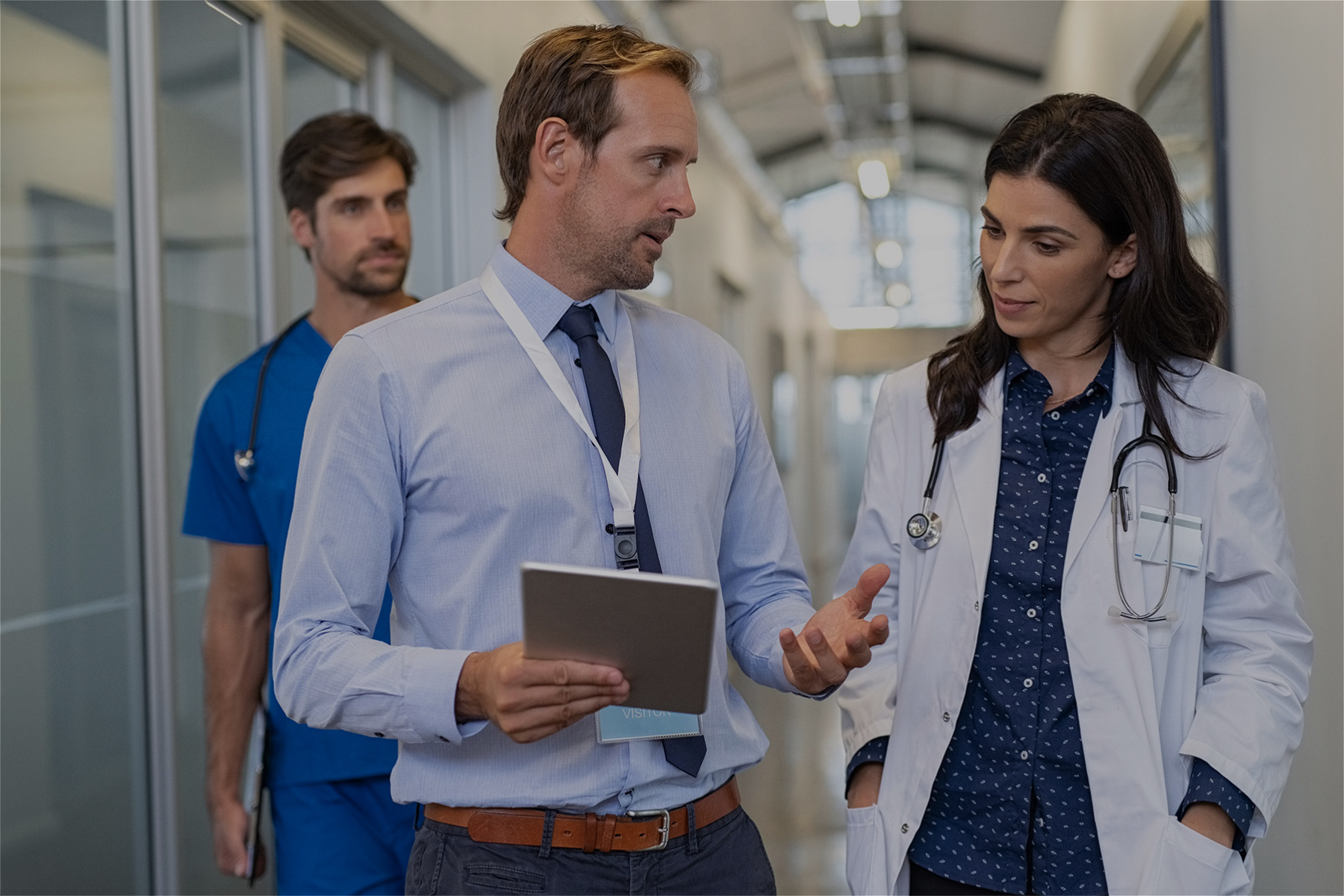 Image of a business man holding a clipboard talking to a doctor with a nurse walking behind them