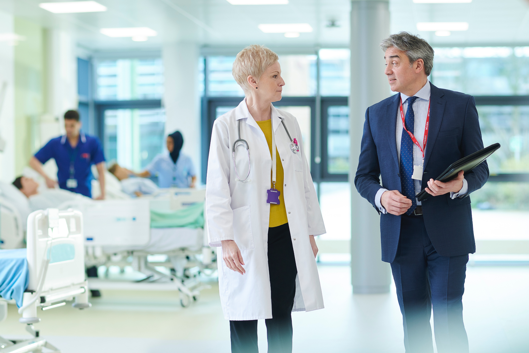 Image of a doctor and a businessman talking to each other while they walk through a hospital ward