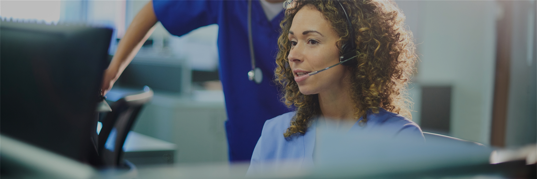 Image of a nurse with headset sitting in front of a computer, and speaking to another nurse standing next to her
