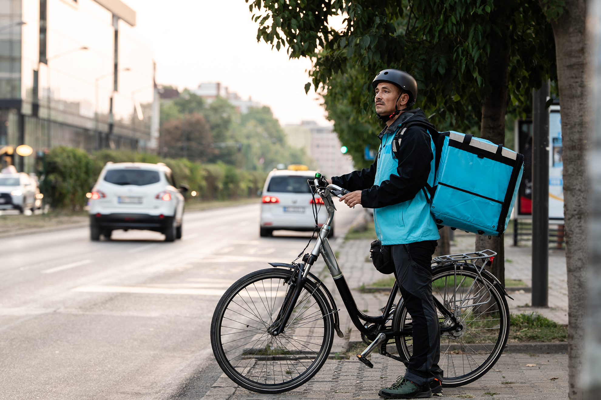 Delivery gig worker with bicycle and thermal backpack waiting to cross the street