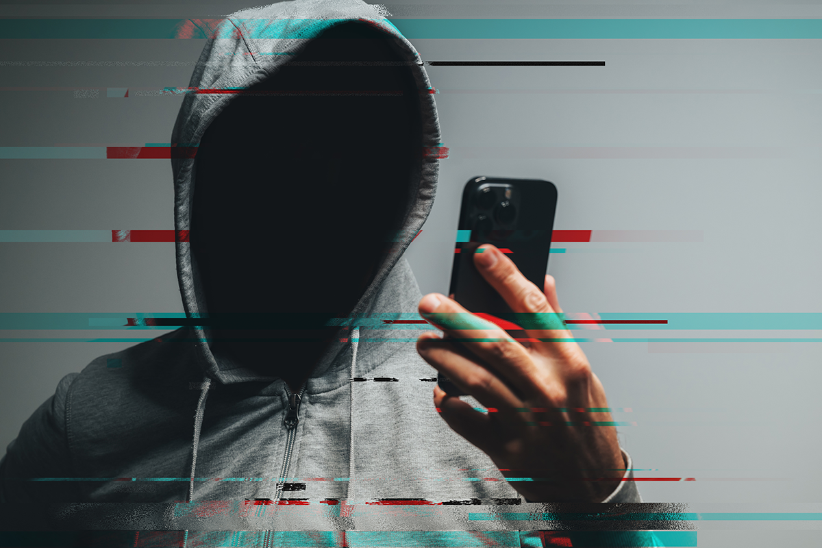 Hooded man with obscured face using smartphone, symbolizing ban evasion, hacker activity, data theft, and cybersecurity threat