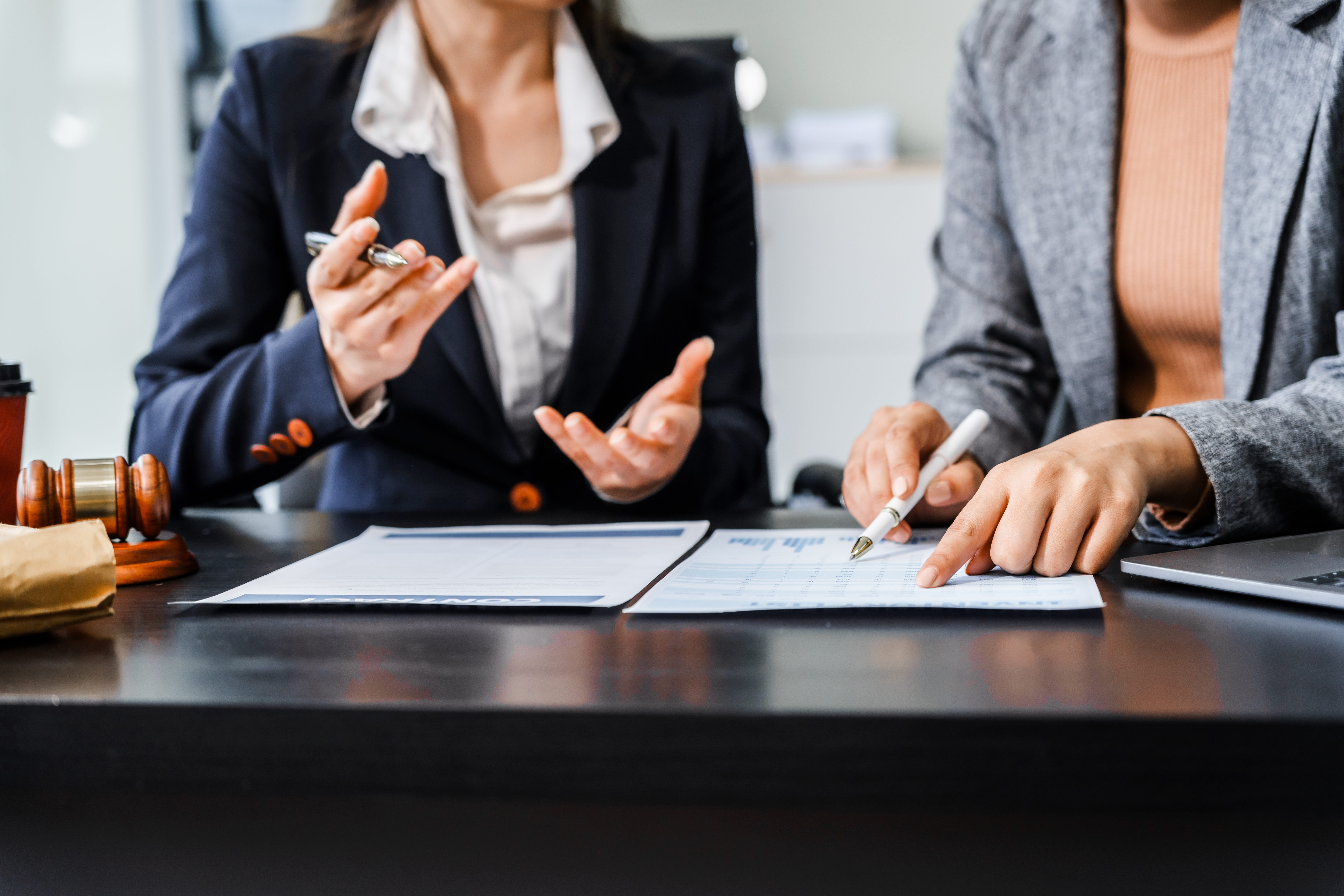 Two female lawyers in business suits meet at wooden desk, shaking hands over contract paper.